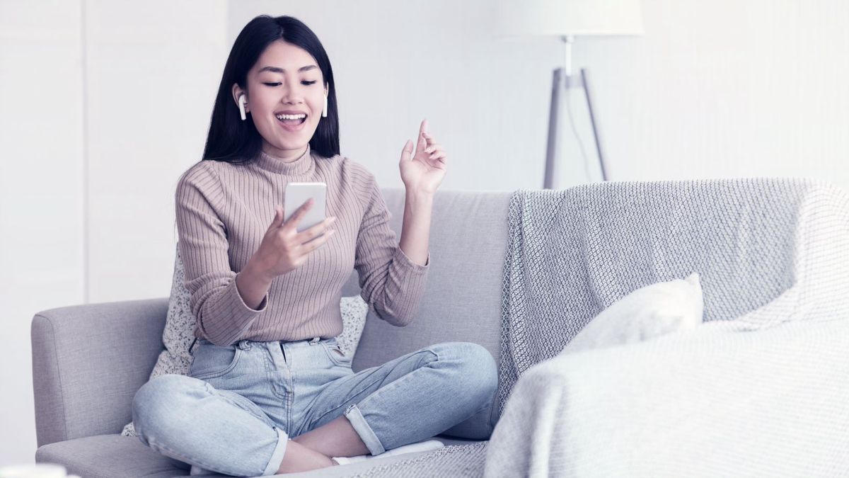 Young woman sitting cross-legged on a couch as she enjoys content from her cell played through earbuds