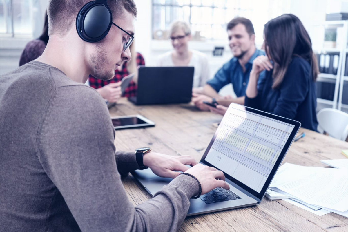 Person wearing noise-cancelling headphones working at a laptop on a table shared with colleagues talking on the same table. 